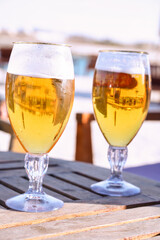 Two glasses of beer on the wooden table with beach club houses reflections. 