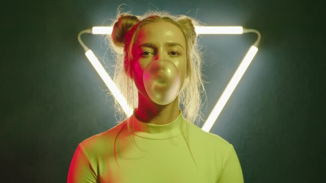 Camera zoom on modern young girl blowing gum bubble in changing colorful light on background with triangular neon lamp