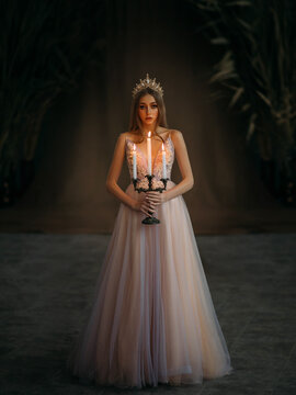 Art photo of medieval girl princess walks in dark gothic room. Woman queen holding candlestick with burning candles in hand. Pink purple dress, long loose blonde hair, gold royal crown. Fairy image