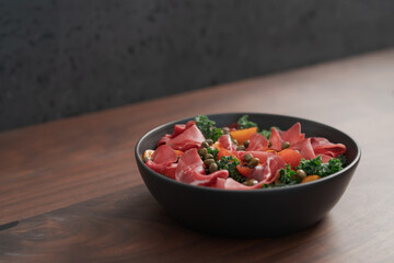 Salad with kale, bresaola and cherry tomatoes decorated with capers in black bowl on walnut wood table
