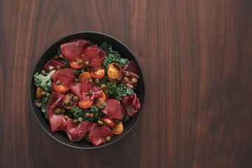 top view of salad with kale, bresaola and cherry tomatoes decorated with capers in black bowl on walnut wood table
