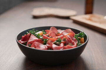 Salad with kale, bresaola and cherry tomatoes decorated with capers in black bowl on walnut wood table