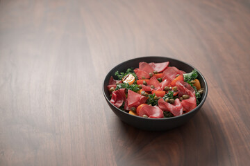 Salad with kale, bresaola and cherry tomatoes decorated with capers in black bowl on walnut wood table