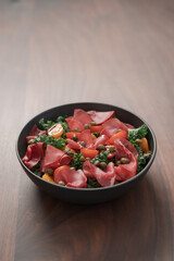 Salad with kale, bresaola and cherry tomatoes decorated with capers in black bowl on walnut wood table