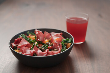Salad with kale, bresaola and cherry tomatoes decorated with capers in black bowl and glass of red drink on walnut wood table