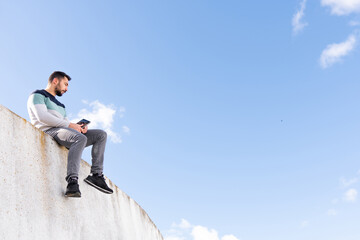 Funny Jump Free caucasian boy with a beard sitting on top of a wall with a tablet making a video call in sunlight outdoors and wearing colorful sweater	
