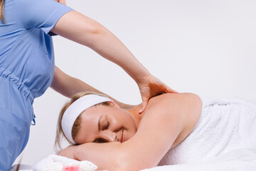 Cropped photo. Hands of the masseur make a reassuring massage to the elder woman who lies wrapped in a white towel with a smile.