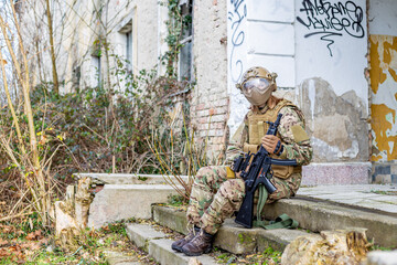 Beautiful girl in military uniform with an airsoft gun sitting on the stairs in front of an abandoned building