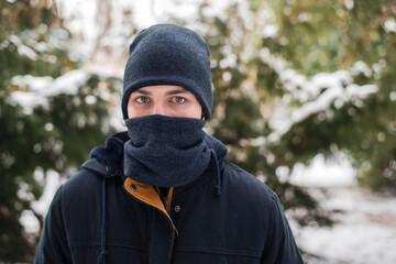 Fototapeta na wymiar A young man in winter clothes and warm hat and scarf covered his whole face, only his eyes are visible, it is very cold outside, snow has fallen, but the guy still spends time with friends outside