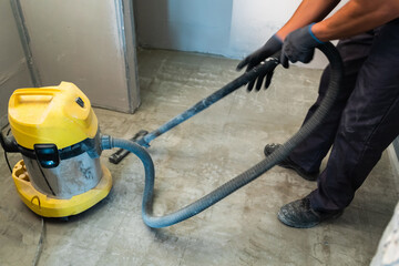 The builder cleans the floor from dust with a construction vacuum cleaner.