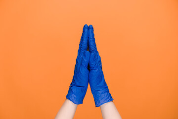 Religion and coronavirus concept. Unrecognizable young female in medical blue gloves holds hands in prayer to god, meditating, praying for health, isolated on orange studio wall with copy space