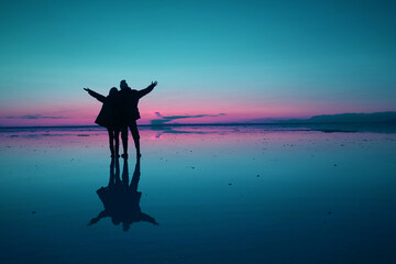 Pop art style blue and pink colored silhouette of happy couple opening arms on the flooding surface of Uyuni salt flats at twilight, Bolivia, South America
