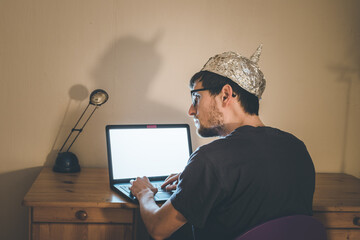 Conspiracy theory concept: young man with aluminum cap searching the internet, sitting lonely in...