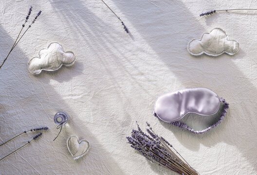 Healthy night sleep creative concept with lavender color. Off white, ivory textile background. Silk sleep mask. Soft toys, clouds and heart shapes. Dry lavender flowers. Natural light, long shadows.