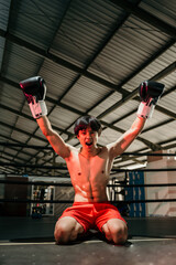Young man looking aggressive in boxing gloves when winning raises both hands sitting on the floor of the boxing ring
