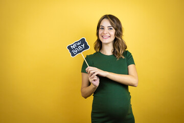 Young beautiful brunette woman pregnant expecting baby over isolated yellow background smiling and holding blackboard with new baby word message