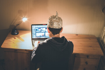 Conspiracy theory concept: young man with aluminum cap searching the internet, sitting lonely in...