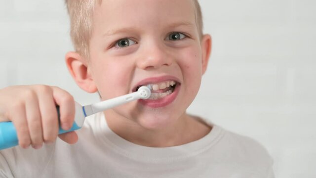 Portrait happy smiling child kid boy brushing teeth with electric toothbrush on white brick background. Health care, dental hygiene.