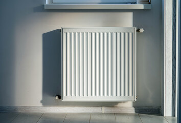 A steel panel heating radiator is placed under the windowsill on a white wall.