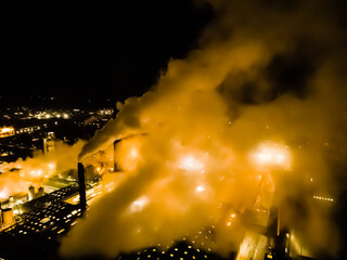 Large factory with thick white smoke coming from the chimneys and bright lights from the factory grounds. Blurred image. 