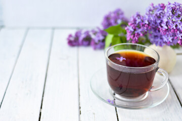 Delicate setting of the morning tea table with lilac flowers, a transparent cup of tea and a saucer and a white vase on a white wooden board. Copy space. Spring breakfast concept