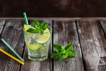 Homemade lemonade in a glass with lime, mint leaves, straw and ice on a dark wooden table. Summer refreshing drink mojito. Selective focus.