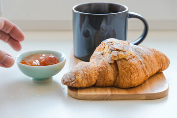 Almond croissant, steaming coffee, or tea. Blurred hand serves apricot jam. Breakfast for one.