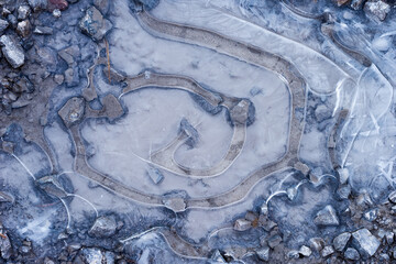 Frozen puddle on a rocky road top view. Snail look pattern made of ice during wintertime. Frozen water with stone embedded