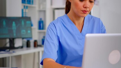Portrait of nurse looking at camera smiling after typing on laptop sitting in hospital office wearing blue uniform. Hospital healthcare worker, medicine clinical assistant health consultant therapist