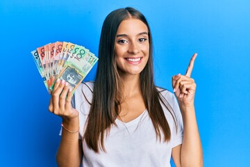 Young hispanic woman holding australian dollars smiling happy pointing with hand and finger to the...