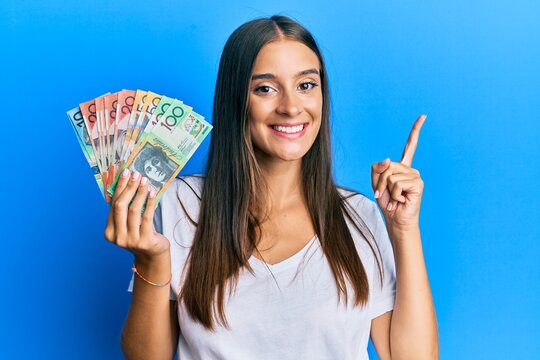Young hispanic woman holding australian dollars smiling happy pointing with hand and finger to the side