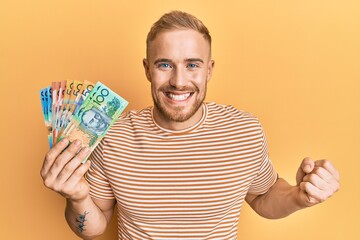 Young caucasian man holding australian dollars screaming proud, celebrating victory and success very excited with raised arm