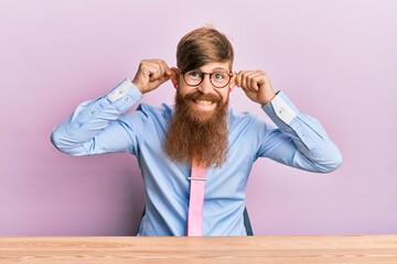 Young irish redhead man wearing business shirt and tie sitting on the table smiling pulling ears...