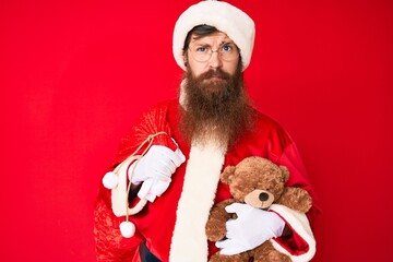 Handsome young red head man with long beard wearing santa claus costume holding teddy bear depressed and worry for distress, crying angry and afraid. sad expression.