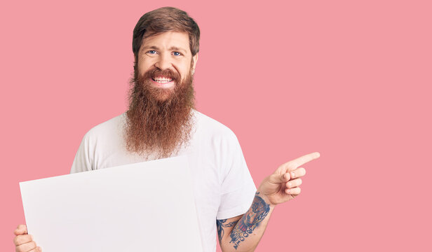 Handsome young red head man with long beard holding blank empty banner smiling happy pointing with hand and finger to the side