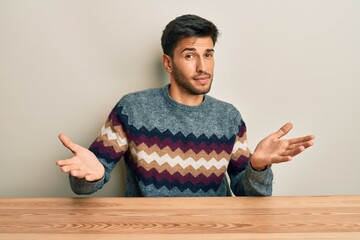 Young handsome man wearing casual sweater sitting on the table clueless and confused expression...