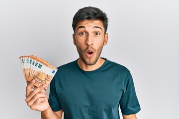 Young handsome man holding bunch of 50 euro banknotes scared and amazed with open mouth for surprise, disbelief face