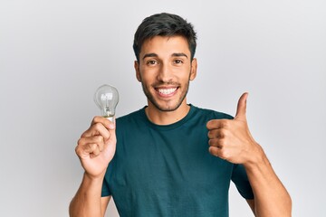 Young handsome man holding lightbulb for inspiration and idea smiling happy and positive, thumb up doing excellent and approval sign