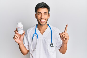 Young handsome man wearing doctor uniform holding presciption pills smiling with an idea or question pointing finger with happy face, number one