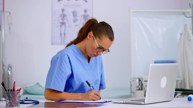 Tired healthcare medical assistant working in hospital researching diagnosing patients health using laptop taking off the glasses having headache. Exhausted therapist in medicine uniform resting eyes