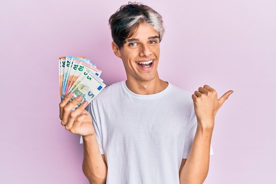 Young hispanic man holding euro banknotes pointing thumb up to the side smiling happy with open mouth