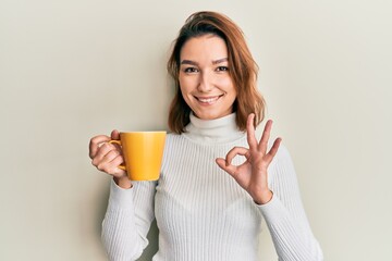 Young caucasian woman holding coffee doing ok sign with fingers, smiling friendly gesturing excellent symbol