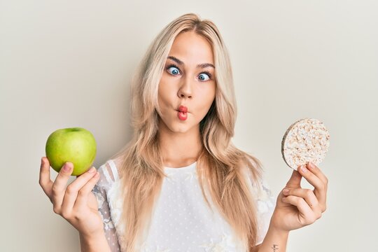 Beautiful caucasian blonde girl holding nachos and healthy green apple making fish face with mouth and squinting eyes, crazy and comical.