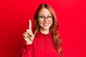 Young beautiful redhead woman wearing casual clothes and glasses over red background showing and...