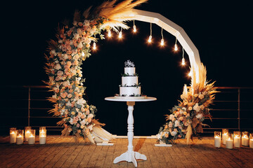 a white cake on a stand against the backdrop of a wedding acre and Edison lamps on, an evening...