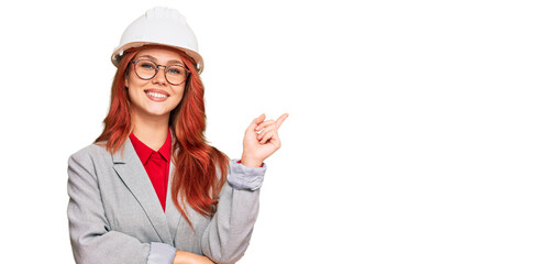 Young redhead woman wearing architect hardhat with a big smile on face, pointing with hand and finger to the side looking at the camera.