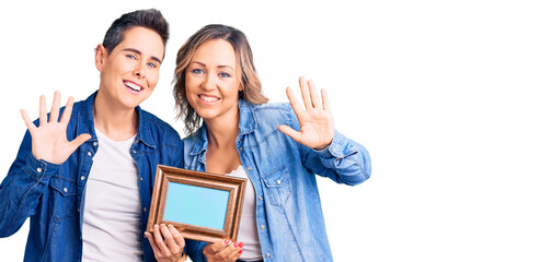 Couple of women holding empty frame waiving saying hello happy and smiling, friendly welcome gesture