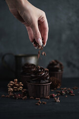 Several muffins or cupcakes with chocolate shaped cream at black table. Festive candle burns on a chocolate cake. A woman's hand crumbles grated chocolate onto a cake
