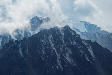 Mountain snowy peaks with clouds on a sunny day. Sunny day high in the mountains, mountain snowy peaks, blue sky, clouds on the tops of mountains, rocks, sharp peaks and steep mountain slopes