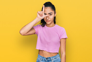 Hispanic teenager girl with dental braces wearing casual clothes making fun of people with fingers on forehead doing loser gesture mocking and insulting.